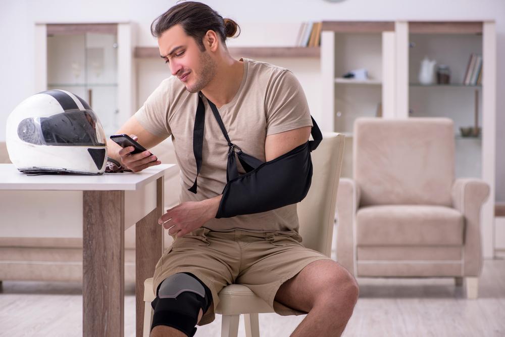 Man with his arm in a sling sits in a chair with a motorcycle helmet on the table next to him as he searches on his phone to ask how to negotiate a higher motorcycle accident settlement.