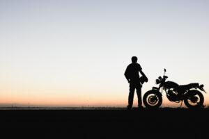 The shadowed silhouette of a motorcyclist holding their helmet while standing next to their bike at sunset, with the rider thinking about the common injuries from motorcycle accidents.