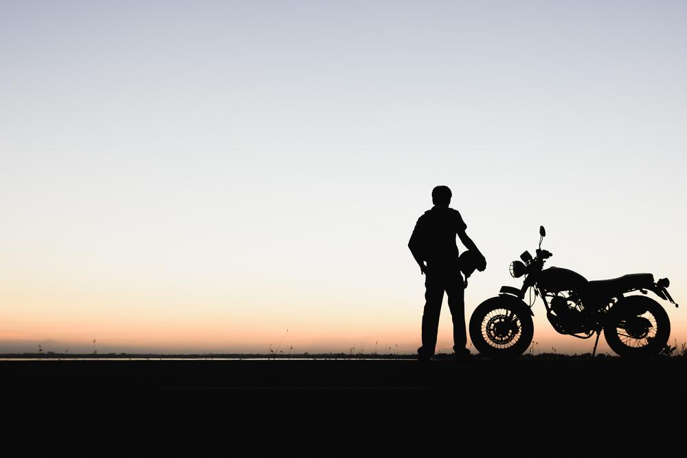 The shadowed silhouette of a motorcyclist holding their helmet while standing next to their bike at sunset, with the rider thinking about the common injuries from motorcycle accidents.