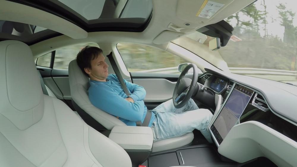 A man relaxing while his Tesla is on autopilot, wondering if he can sue Tesla for a crash involving autopilot.